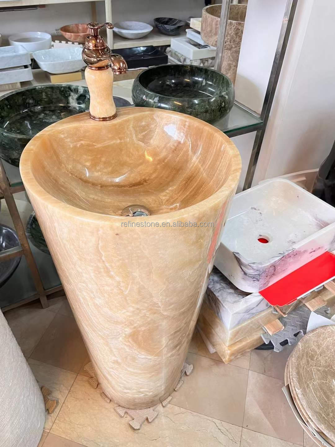 Luxury tall cylindrical standing marble basin sink by Lise Luxury with a bronze faucet, surrounded by various stone bathroom fixtures with customized options.