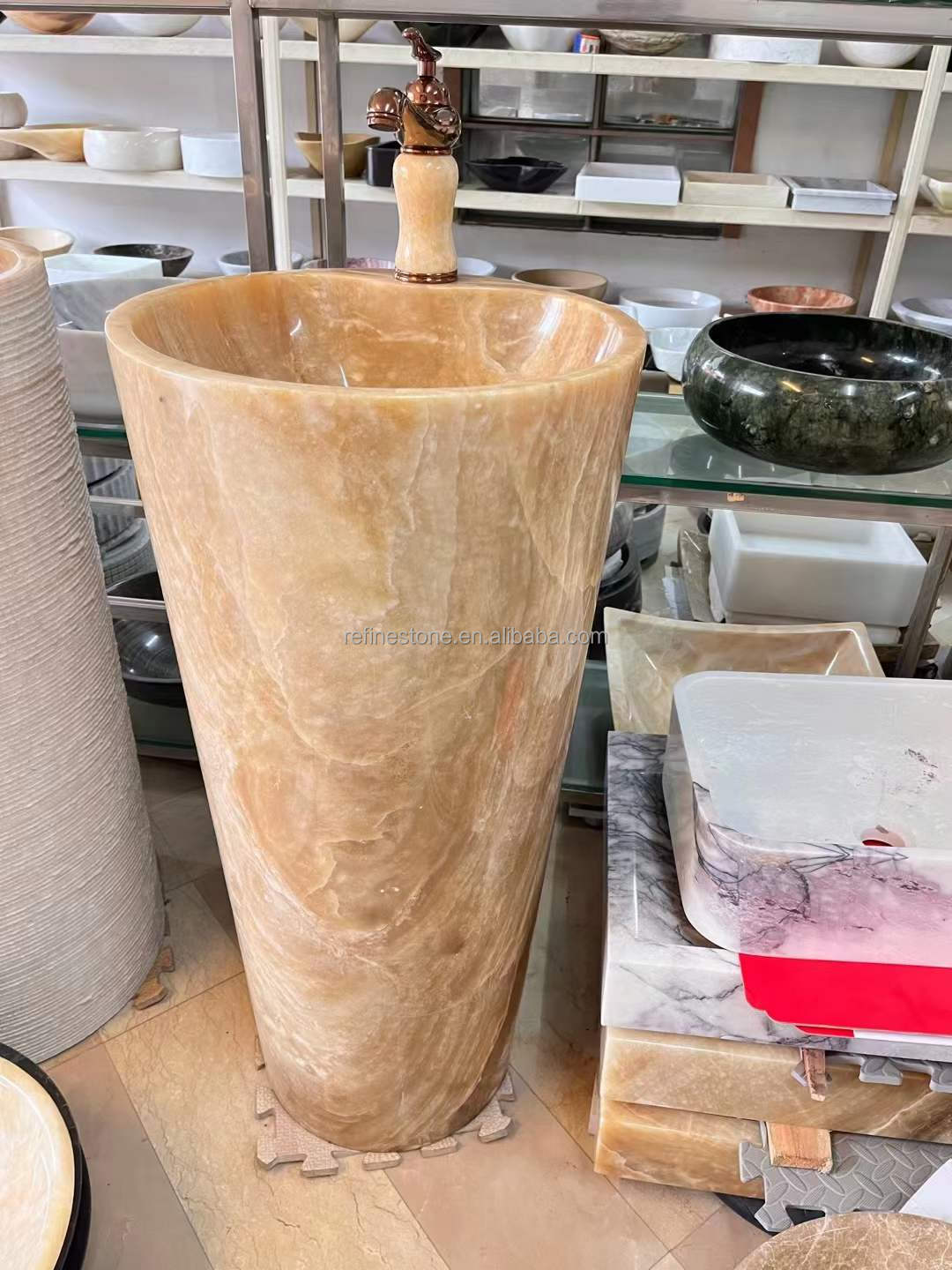 Luxury tall cylindrical standing marble basin sink by Lise Luxury with a bronze faucet, surrounded by various stone bathroom fixtures with customized options.