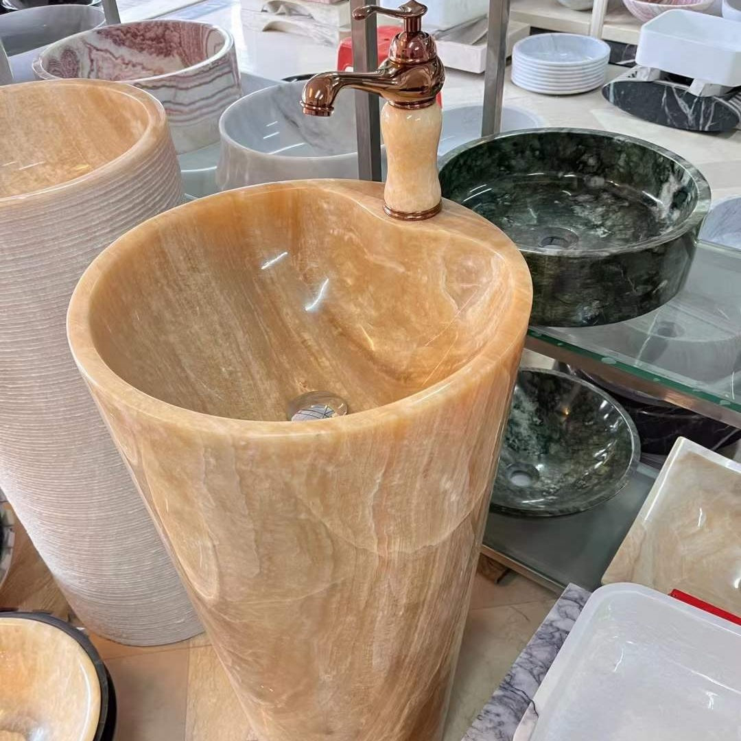 Luxury tall cylindrical standing marble basin sink by Lise Luxury with a bronze faucet, surrounded by various stone bathroom fixtures with customized options.