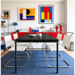[color: ivory] A modern dining room scene with a black rectangular table and six white cantilever chairs with chrome S-shaped legs, set on a blue rug in a brightly lit room with colorful abstract wall art and red cabinetry.