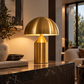 [color: gold] A modern mushroom-shaped table lamp in polished brass sits on a dark marble console table in a stylishly decorated living room.