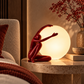 Red sculptural table lamp featuring a kneeling human figure with one arm raised beside a glowing white orb