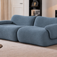 A modern, low-profile loveseat sofa upholstered in a textured slate blue fabric, set in a contemporary living room with a beige rug, wooden floor, and dark shelving.