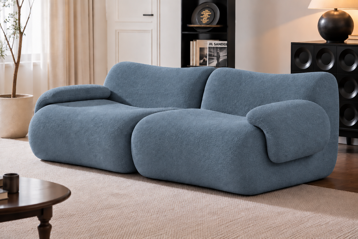 A modern, low-profile loveseat sofa upholstered in a textured slate blue fabric, set in a contemporary living room with a beige rug, wooden floor, and dark shelving.