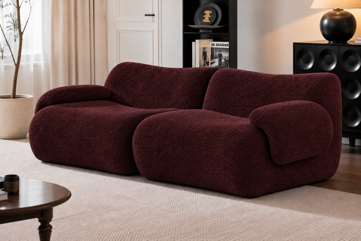 A modern, low-profile sofa upholstered in a deep burgundy bouclé fabric, set in a styled living room with a beige rug, a potted plant, and a black sideboard.
