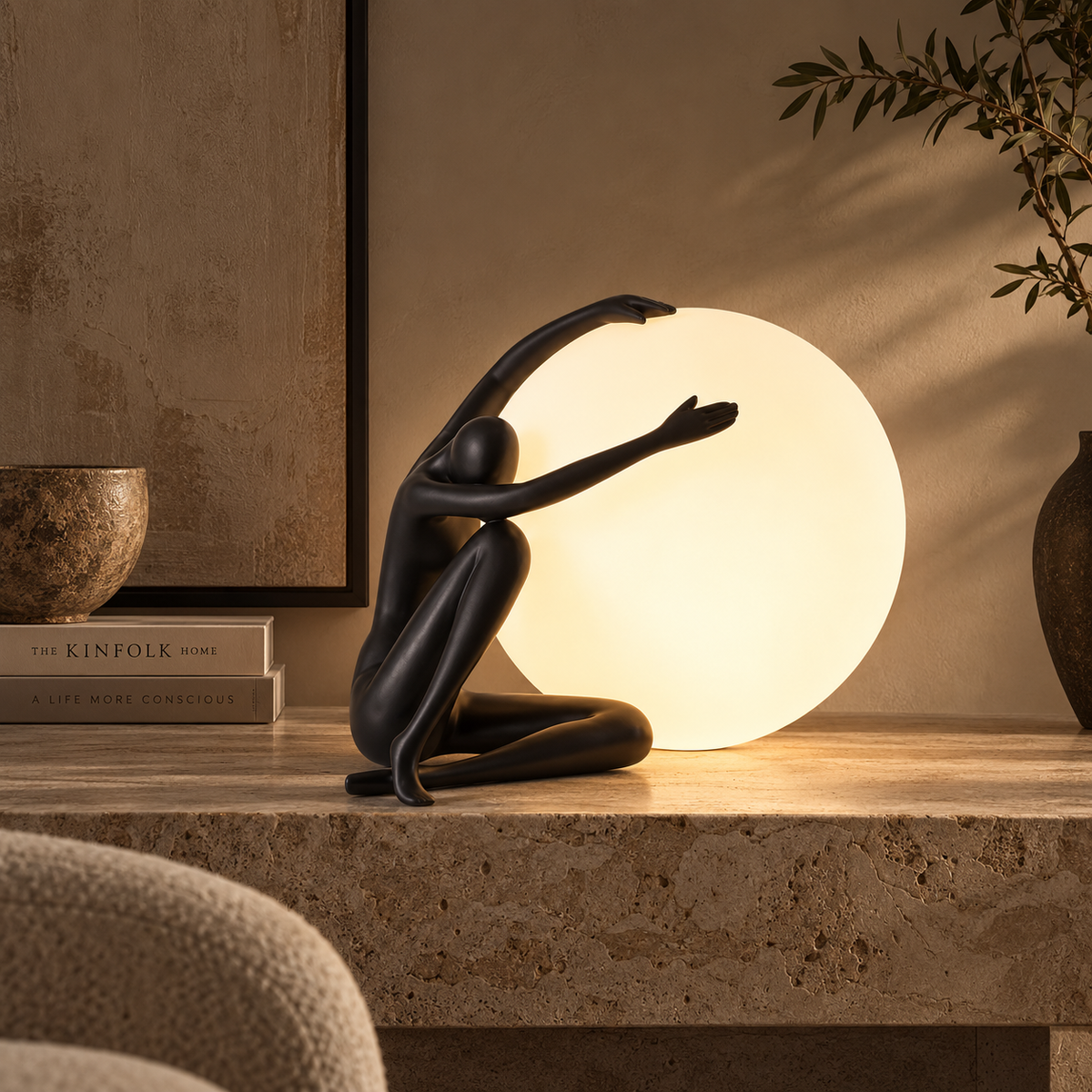 A black sculptural figurine of a kneeling human figure with arms raised, placed beside a glowing white spherical table lamp on a stone surface in a warmly lit interior setting.
