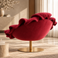 [color:pink] A luxurious deep red velvet lounge chair with dramatic ruffled detailing on the backrest, mounted on a brushed gold metal pedestal base, set in a warm, sunlit modern room.