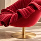 [color:pink] A close-up of a luxurious red velvet swivel chair with dramatic pleated detailing and a brushed gold metal pedestal base, set against a neutral beige background.