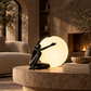 Black figurative table lamp with a sculptural human figure reaching toward a glowing white globe in a modern interior