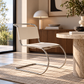 [color: ivory] A modern cantilever chair with a cream upholstered seat and backrest and a curved chrome metal base, shown in a stylishly decorated living room next to a marble side table.