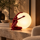 Red figurative table lamp with a seated human figure beside a glowing white spherical shade