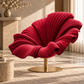 [color:pink] A modern shell-shaped armchair in deep red pleated velvet with a gold pedestal base, set in a warm, sunlit contemporary living room.