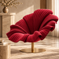 [color:pink] A modern red upholstered accent chair with a wavy, shell-like backrest and a round gold metal base, shown in a warm, sunlit interior setting.