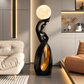 A modern abstract floor lamp with a glowing spherical moon-like bulb, a black sculptural body, and a gold interior accent, placed in a contemporary living room beside a sofa and chair.
