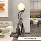 A modern abstract floor lamp with a glowing spherical top and a sculptural metallic base, shown in a furnished living room. A text overlay at the bottom reads 'SIZE: 24*24*82CM'.
