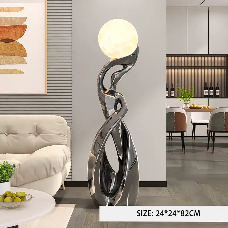 A modern abstract floor lamp with a glowing spherical top and a sculptural metallic base, shown in a furnished living room. A text overlay at the bottom reads 'SIZE: 24*24*82CM'.