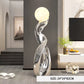 A modern abstract floor lamp with a glowing spherical top and a reflective metallic sculptural base, shown in a styled living room interior. A size label at the bottom reads 24*24*82CM.