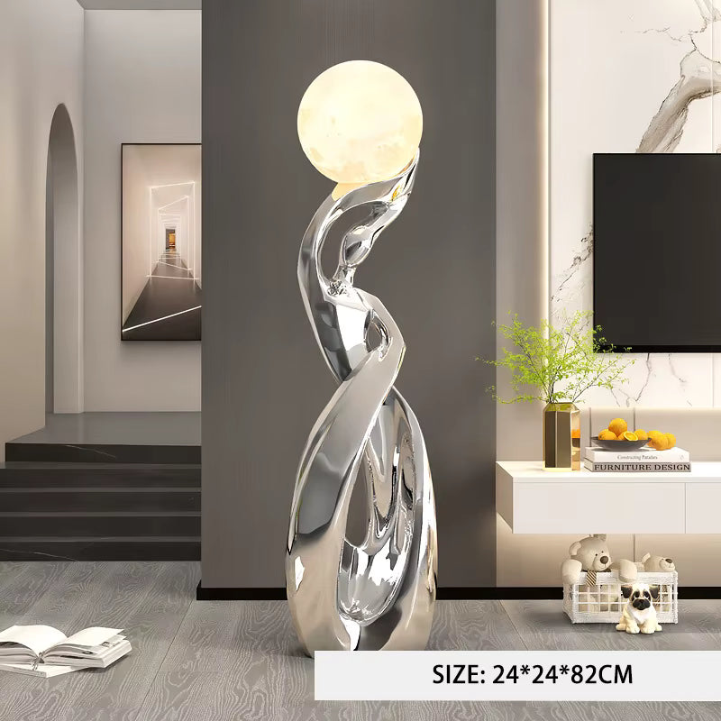 A modern abstract floor lamp with a glowing spherical top and a reflective metallic sculptural base, shown in a styled living room interior. A size label at the bottom reads 24*24*82CM.