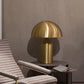 [color: gold]  Gold table lamp on a side table next to a textured chair against a gray wall.