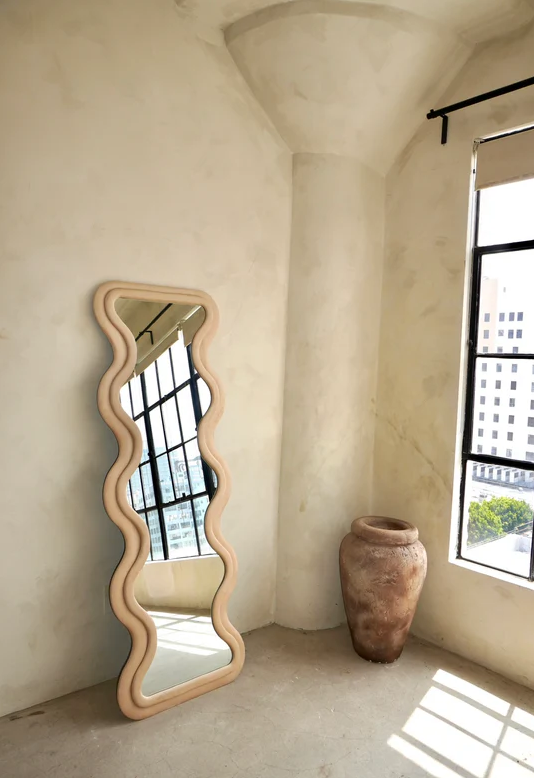 [color:rose]A tall, wavy-edged floor rose colored mirror with a light natural wood frame leans against a textured beige plaster wall in a sunlit room. A large terracotta vase sits near a window on the right.
