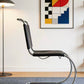 [color: noir] A modern cantilever chair with a black leather seat and backrest and a curved chrome base, shown in a styled room setting with a blue rug, floor lamp, and abstract art on the wall.