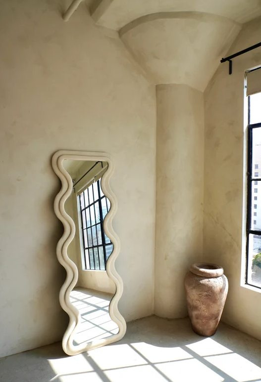 [color:ivory] A tall floor-length ivory color mirror with a wavy, scalloped cream-colored frame leaning against a textured beige plaster wall in a sunlit room. A large rustic terracotta vase sits on the floor near a window, and natural light casts shadows across the room.