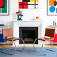 [color: cognac] Two modern cantilever armchairs with brown leather seats and chrome steel frames, positioned symmetrically in front of a white fireplace in a brightly lit room with colorful abstract wall art.