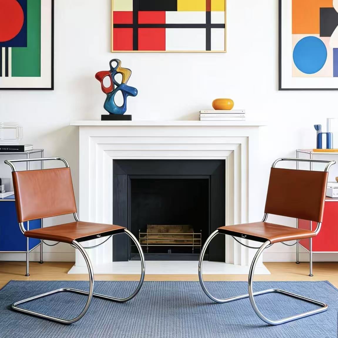 [color: cognac] Two modern cantilever armchairs with brown leather seats and chrome steel frames, positioned symmetrically in front of a white fireplace in a brightly lit room with colorful abstract wall art.