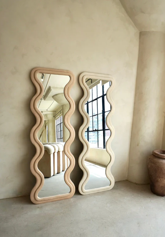 [color:rose] Two tall wavy-edged mirrors in colors ivory and rose with light natural wood frames leaning against a textured beige plaster wall in a minimalist interior setting.