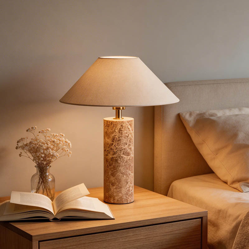 A table lamp with a beige fabric drum shade and a cylindrical marble base sits on a wooden bedside table next to an open book and a small vase of dried flowers, with a bed in the background.