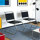 [color: noir] Two modern cantilever chairs with black mesh seats and backs and chrome curved metal bases, placed on a blue rug in a styled room with framed wall art and a floor lamp.