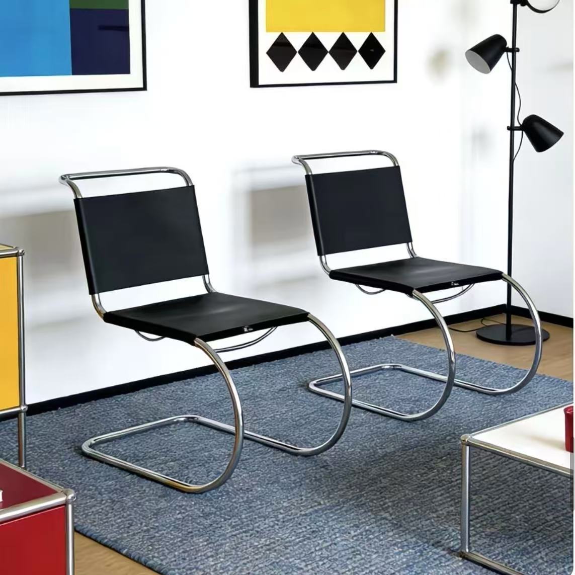 [color: noir] Two modern cantilever chairs with black mesh seats and backs and chrome curved metal bases, placed on a blue rug in a styled room with framed wall art and a floor lamp.