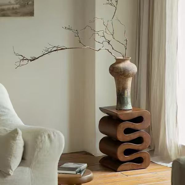 [color: brown] A rustic ceramic vase with a textured earthy brown and green glaze sits on a sculptural wavy wooden side table. The table is styled in a cozy, sunlit room next to a cream-colored armchair and draped curtains.