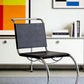 [color: noir] A modern cantilever chair with a black upholstered seat and backrest and a curved chrome metal base, shown in a styled interior setting with a white cabinet, books, and colorful wall art.