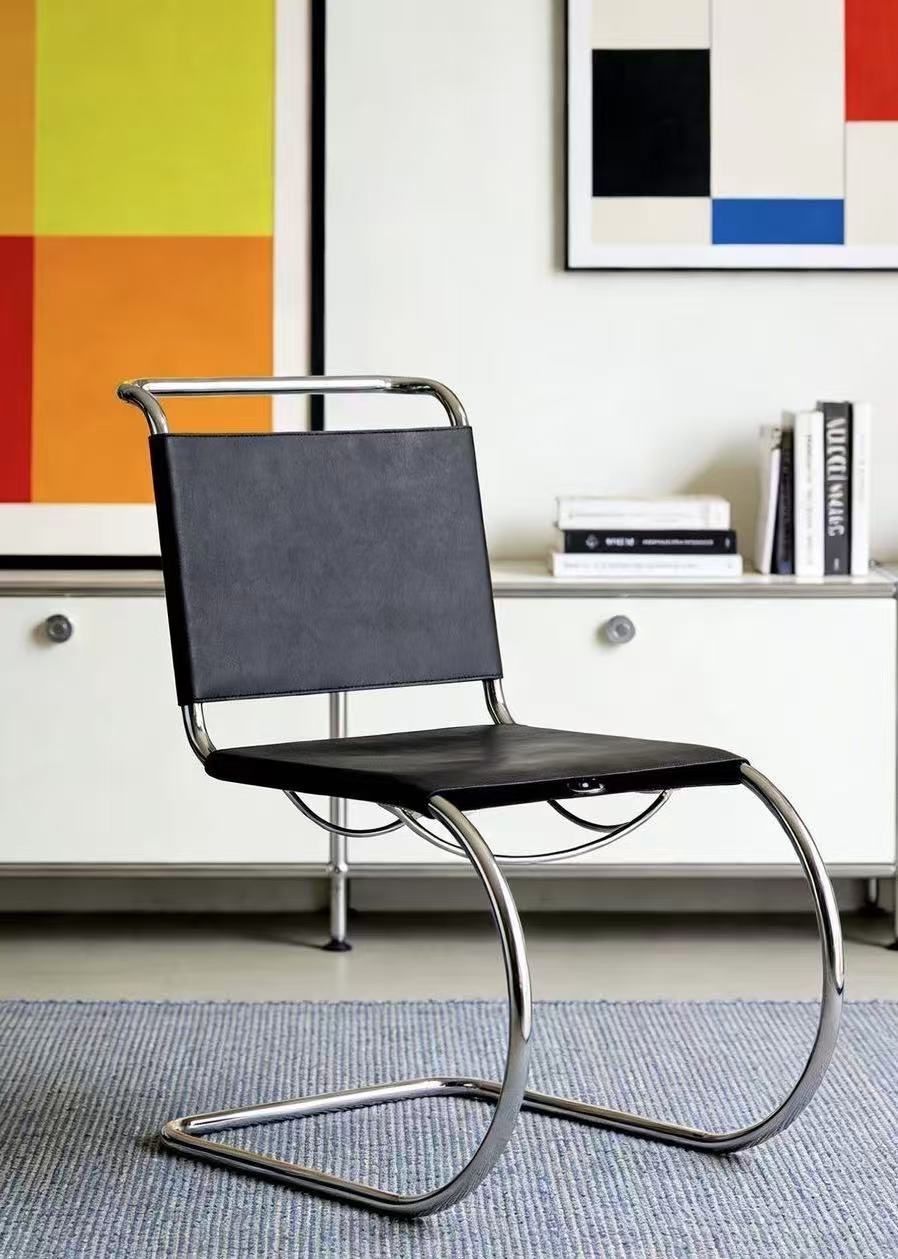 [color: noir] A modern cantilever chair with a black upholstered seat and backrest and a curved chrome metal base, shown in a styled interior setting with a white cabinet, books, and colorful wall art.