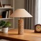 A table lamp with a beige fabric drum shade and a cylindrical marble base sits on a wooden desk in a home office setting, flanked by a potted plant and a wooden clock.