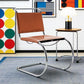 [color: cognac] A cantilever chair with a brown leather seat and backrest and a curved chrome steel base, shown in a styled room setting next to a side table with a lamp.