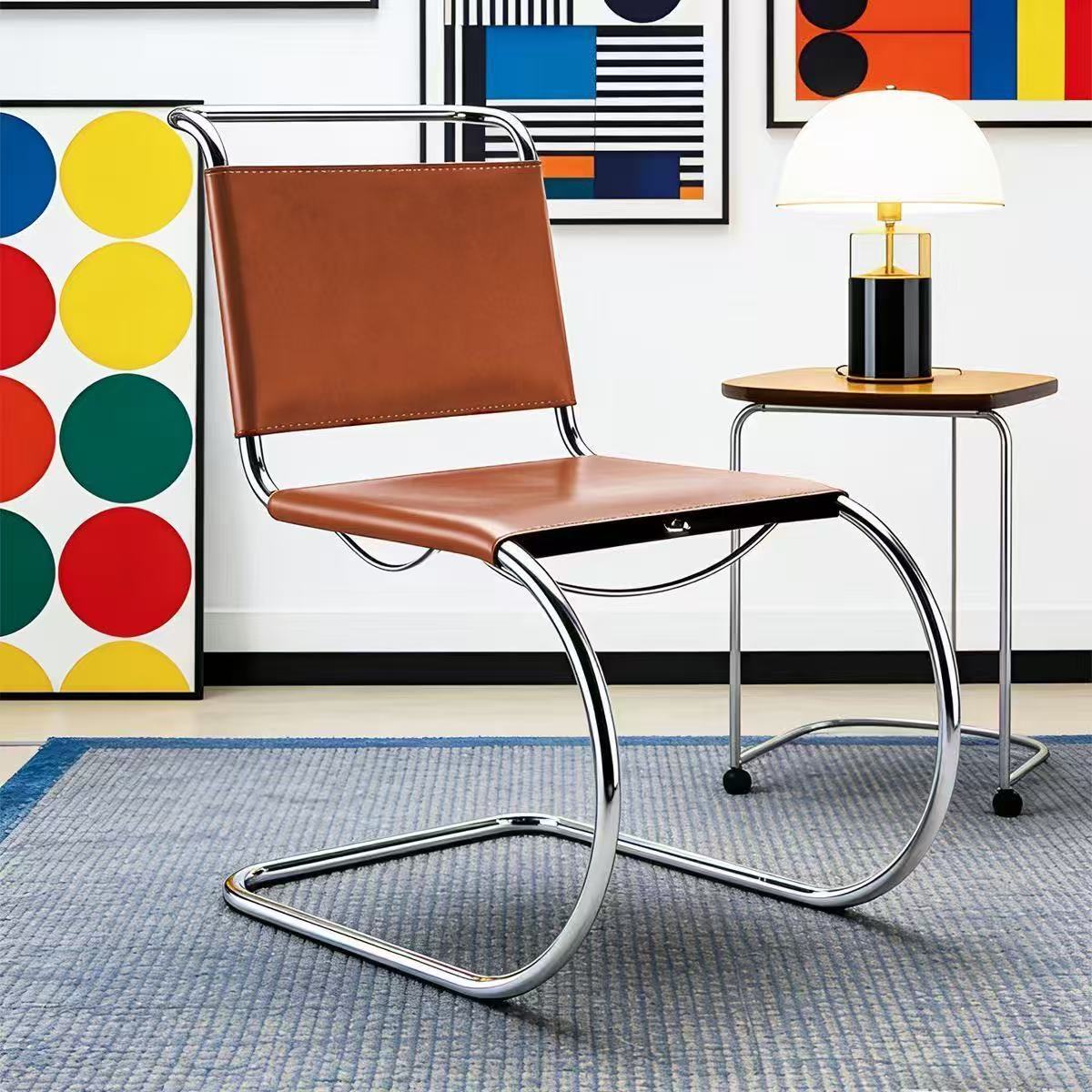 [color: cognac] A cantilever chair with a brown leather seat and backrest and a curved chrome steel base, shown in a styled room setting next to a side table with a lamp.
