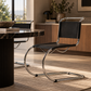 [color: noir] A modern cantilever chair with a black leather seat and backrest and a curved polished chrome base, positioned in a stylishly decorated room next to a marble-top dining table.