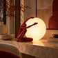 A sculptural table lamp featuring a red crouching human figure reaching toward a large glowing white globe, placed on a stone console table in a warmly lit interior setting.