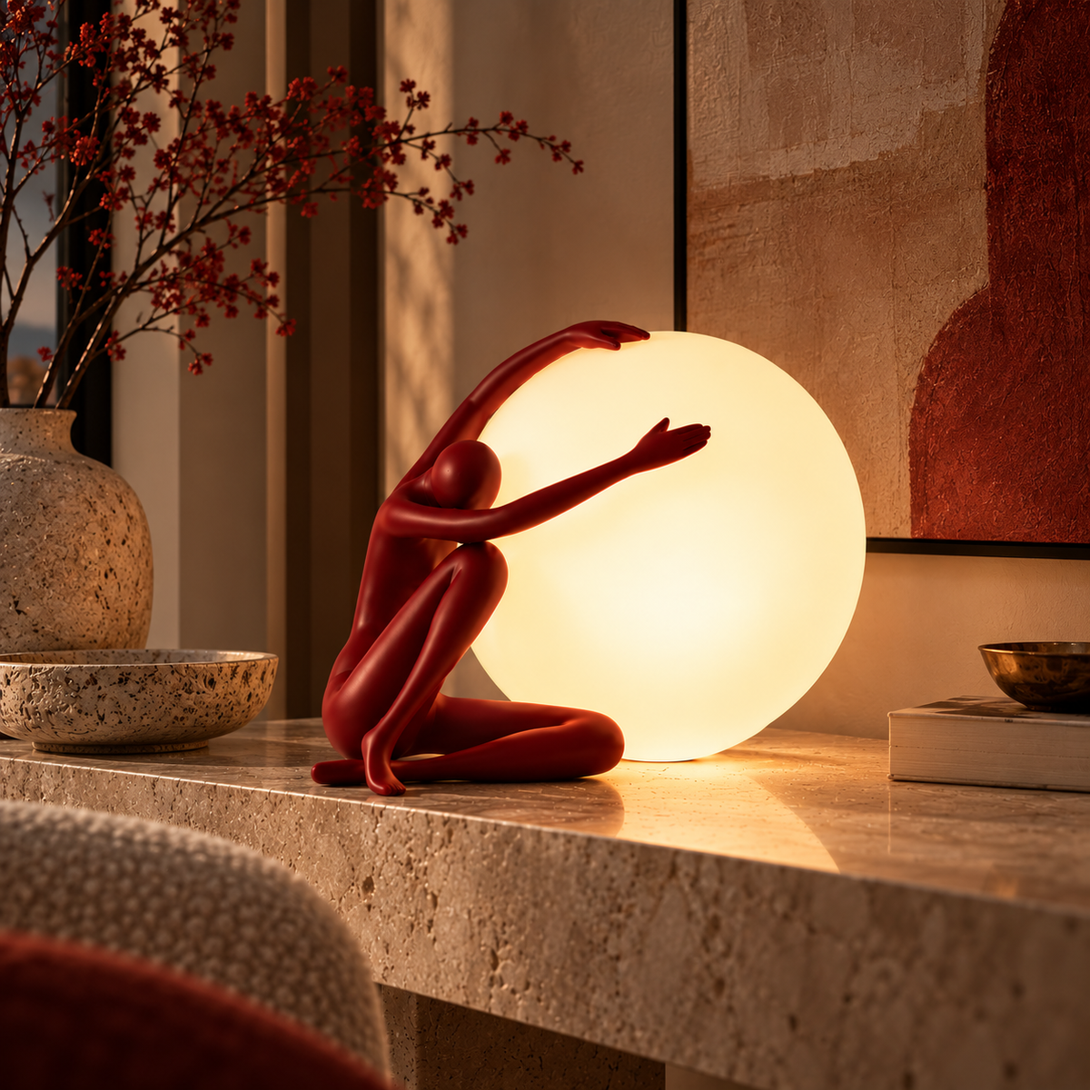 A sculptural table lamp featuring a red crouching human figure reaching toward a large glowing white globe, placed on a stone console table in a warmly lit interior setting.