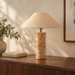 A table lamp with a beige fabric drum shade and a cylindrical beige stone base sits on a dark wood console table in a warm, sunlit room. A potted plant and a book are visible on the left, and a framed print is on the right.