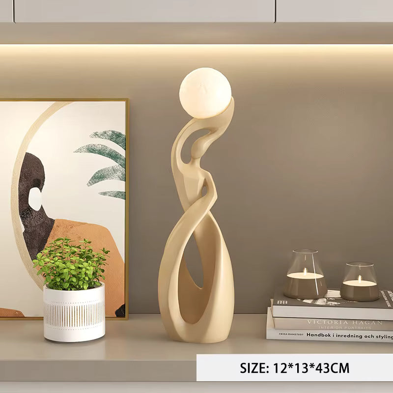 A modern abstract floor lamp with a cream-colored sculptural base and a glowing white spherical shade, displayed on a shelf next to a potted plant and books.