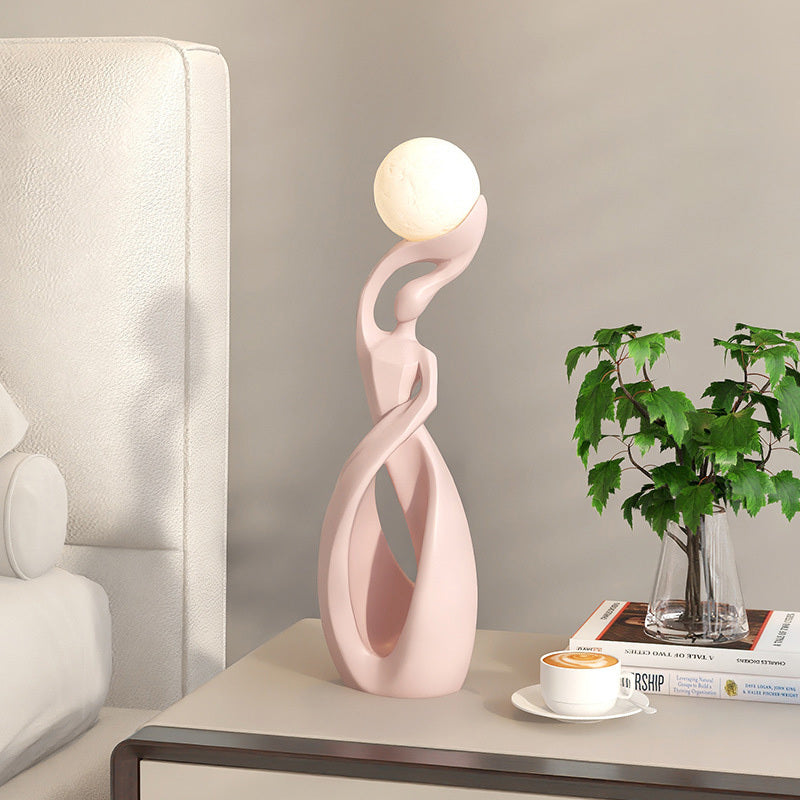 A modern sculptural table lamp in a light pink color with a glowing white spherical shade, styled on a bedside table next to a white leather headboard, a small plant in a glass vase, and a cup of coffee.