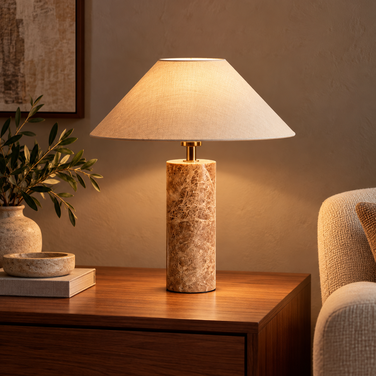 A table lamp with a warm beige fabric drum shade and a cylindrical marble base in beige and brown tones, lit and placed on a wooden side table in a cozy living room setting.