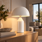 [color: white] A modern table lamp with a glossy white dome-shaped shade and a cylindrical white base sits on a wooden console table in a warm, softly lit living room.
