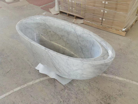 A luxury oval Lise Luxury grey-white marble bathtub on a concrete floor with wooden crates in the background.